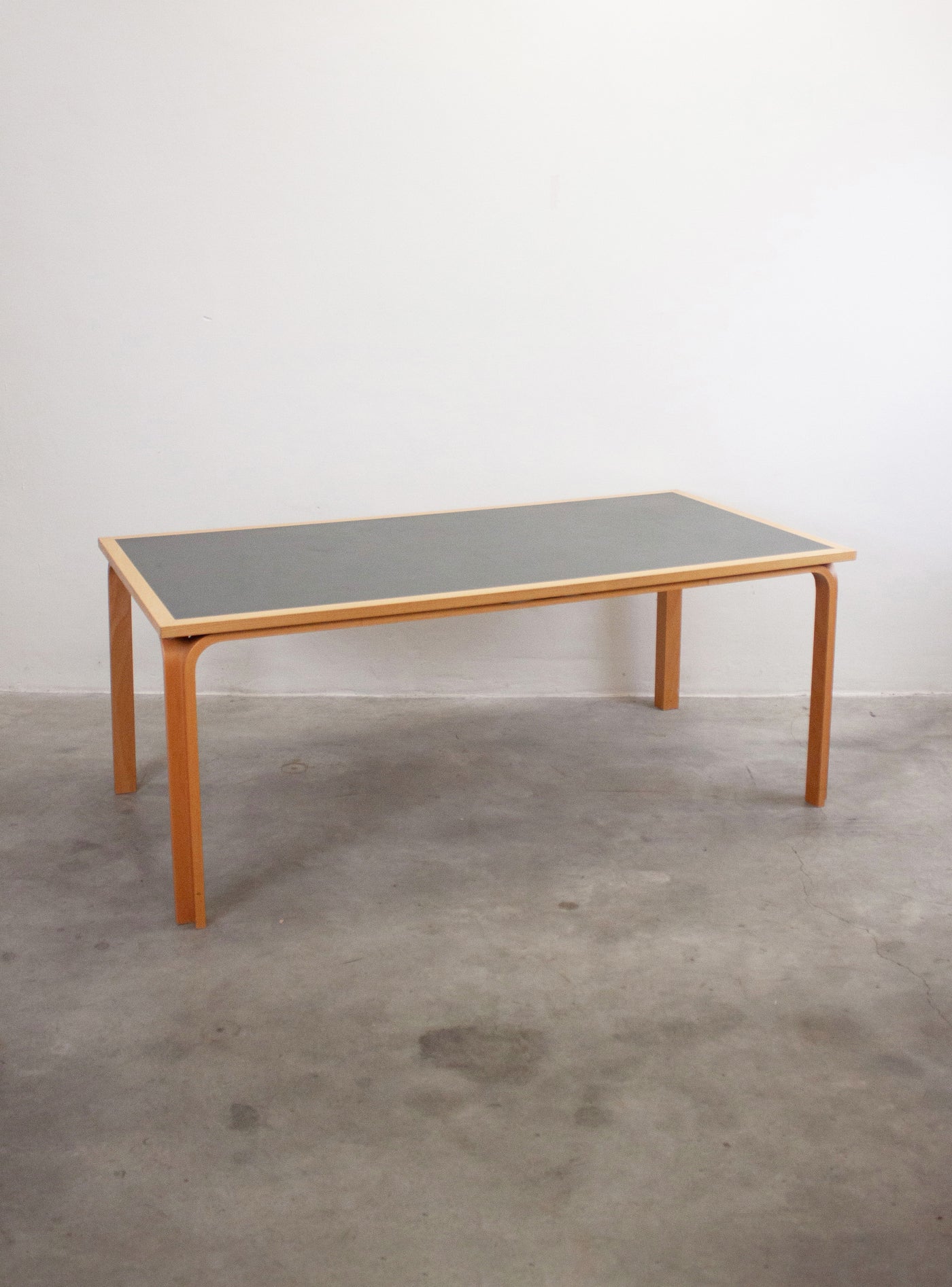 Vista 3/4 del tavolo vintage Magnus Olesen anni '50 — piano in linoleum grigio con cornice in legno chiaro, gambe in legno curvato