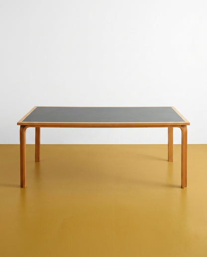 Vista frontale ambientata del tavolo Magnus Olesen — piano bicolore linoleum e legno chiaro, gambe curve