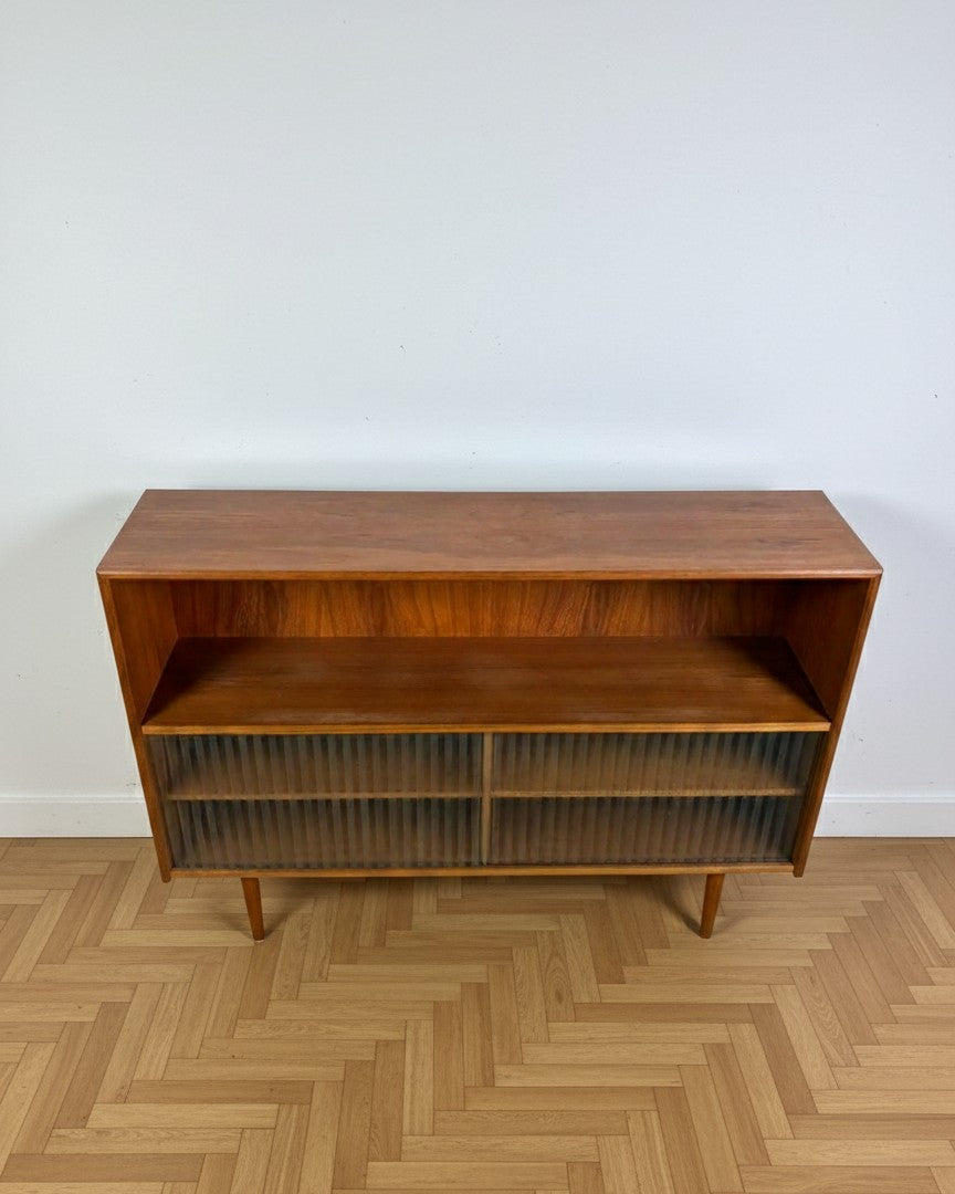 Libreria bassa danese anni 50 in teak, vista frontale su sfondo neutro, ante scorrevoli in vetro plissettato rigato e gambe affusolate coniche vista dall'alto